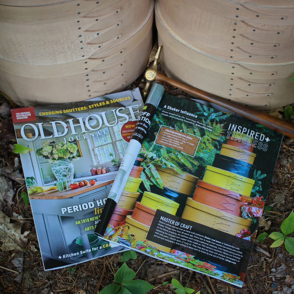 Old House Journal, September/October 2026, open to the full-page feature "A Shaker Influence," resting on the forest floor beside a brass tack hammer and two large unfinished Shaker oval boxes.