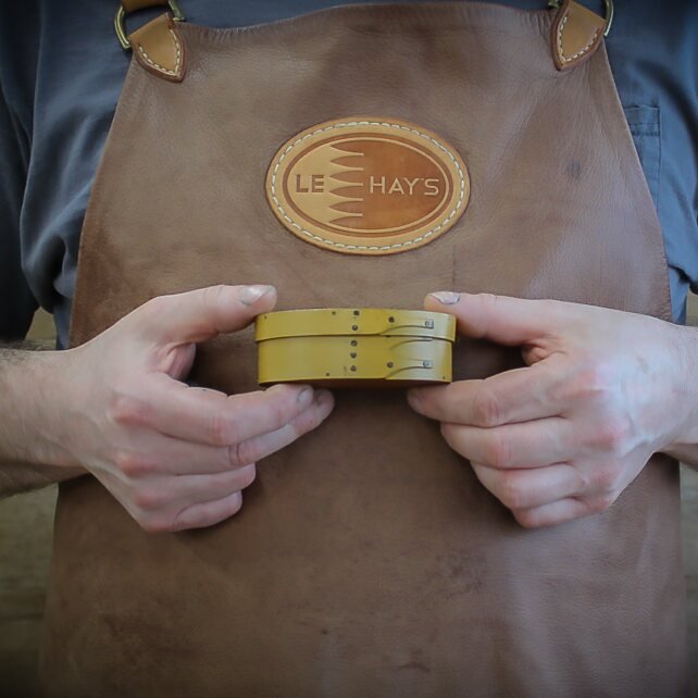 Handcrafted wooden oval shaker box held by a person in a leather apron, perfect for sewing and needlework storage.