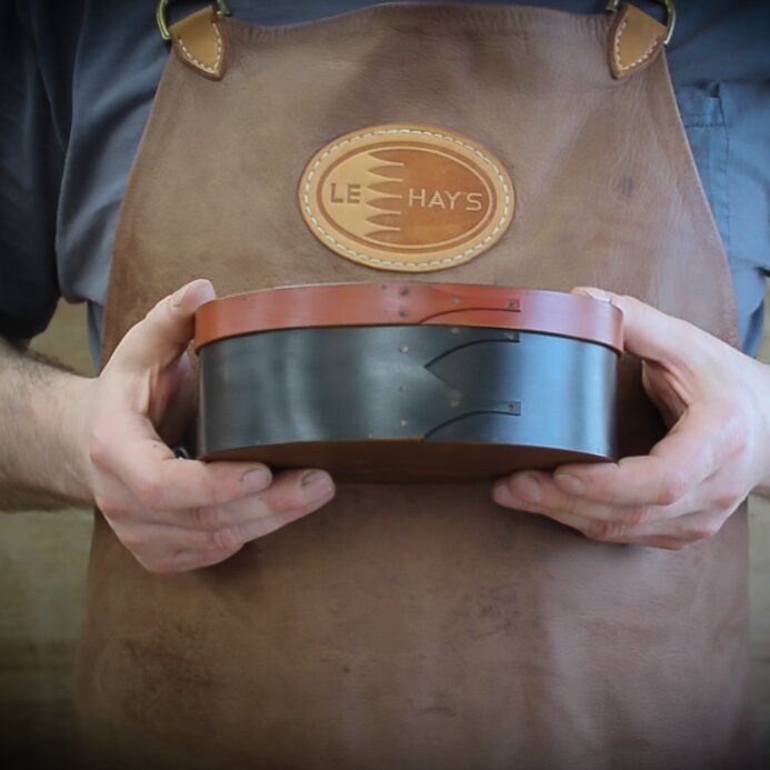 Handcrafted wooden oval sewing box from Maine, ideal for needlework and storage, held by artisan in leather apron.